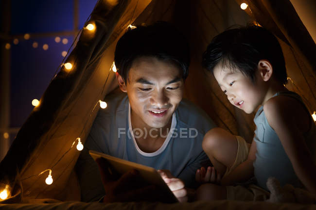 Щасливий азіатський батько і дошкільник син дивиться щось на планшеті в наметі вдома — стокове фото