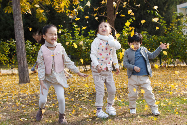Счастливые мальчики и девочки, играющие вместе с осенними листьями в парке — стоковое фото