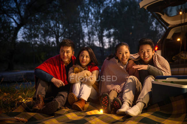 Vier lächelnde asiatische Freunde sitzen in der Nähe des Autos und schauen in die Kamera im herbstlichen Abendwald — Stockfoto