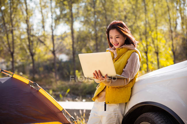 Alegre asiático mujer holding laptop y apoyándose en coche en otoñal bosque - foto de stock