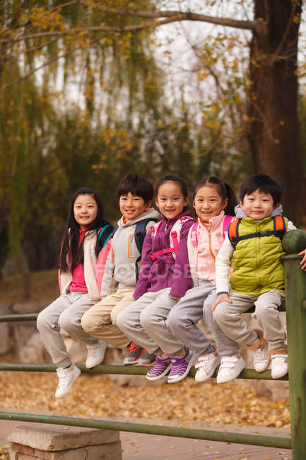 Five adorable smiling asian kids sitting on fence and looking at camera in autumnal park — Stock Photo