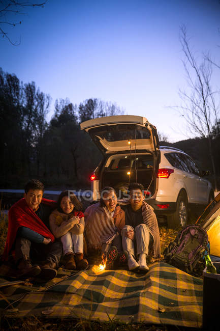 Quatre souriant asiatique amis assis près de voiture et regarder caméra dans automne soirée forêt — Photo de stock