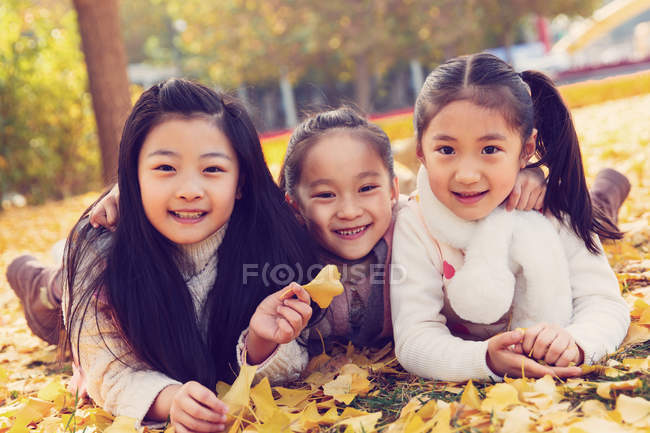 Three adorable asian kids lying on yellow foliage and holding leaves in autumnal park — Stock Photo