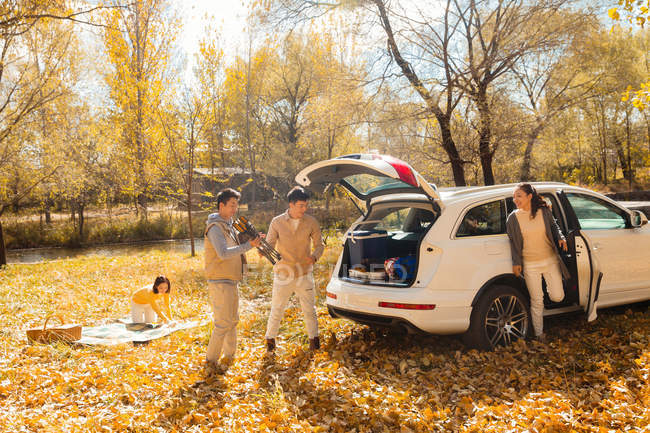 Male and female asian friends taking stuff for picnic from car in autumnal forest — Stock Photo