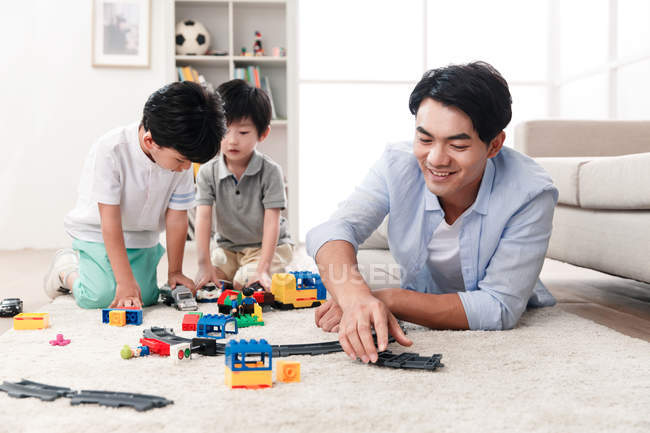 Happy asian father and sons playing with toy railway station on floor at home — Stock Photo