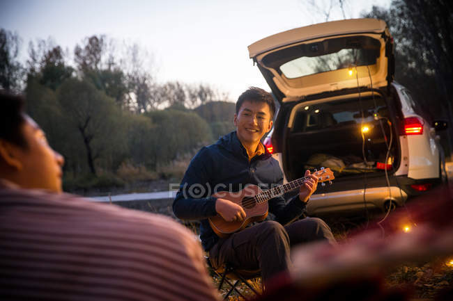 Glücklicher asiatischer Mann spielt Ukulele für Freund im herbstlichen Wald — Stockfoto