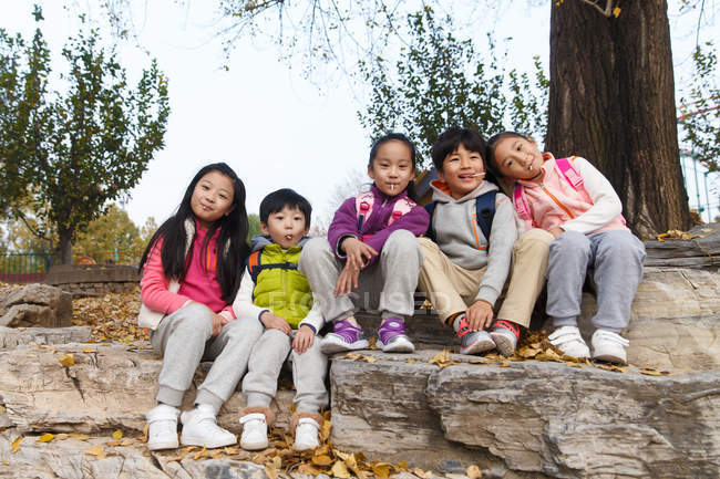 Five adorable asian kids sitting on stones and looking at camera in autumnal park — Stock Photo