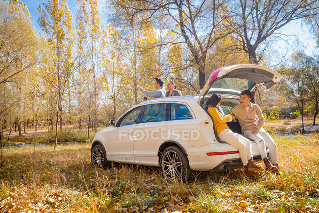 Male and female asian friends sitting in car in autumnal forest — Stock Photo