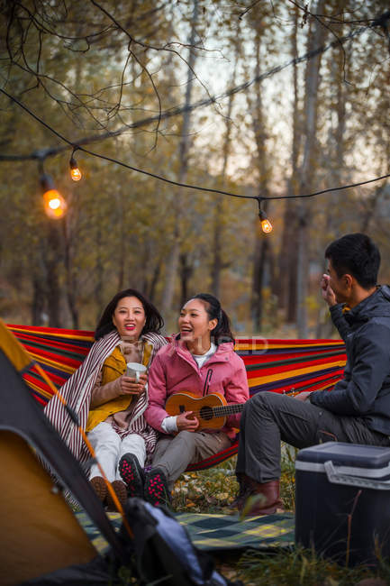 Smiling asian friends having fun with ukulele on hammock in autumnal forest — Stock Photo