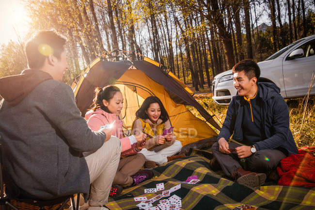 Four asian friends playing cards on blanket near tent in autumnal forest — Stock Photo