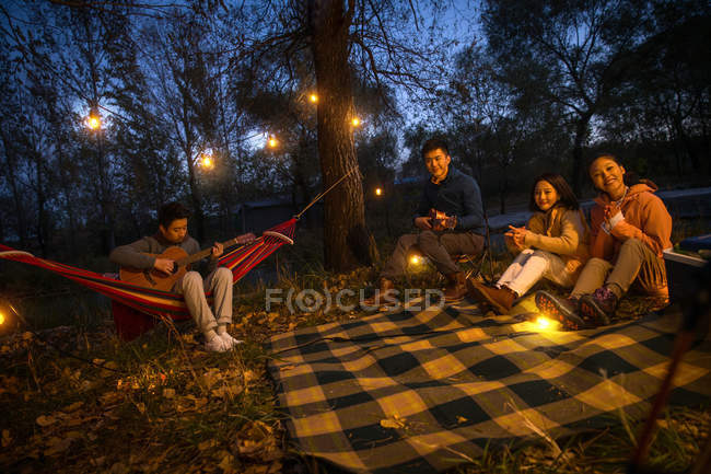 Hombre jugando guitarra para tres asiático amigos en camping en otoñal noche bosque - foto de stock