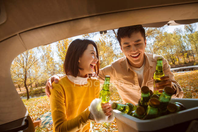 Feliz asiático pareja tomando cerveza botellas de coche en otoñal bosque - foto de stock