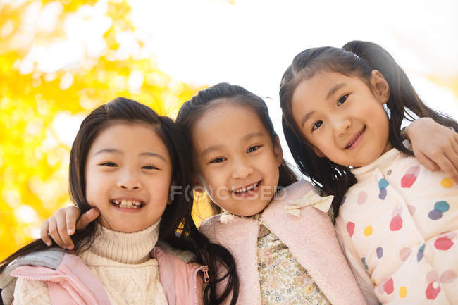 Low angle view of three adorable smiling asian kids hugging in autumnal park and looking at camera — Stock Photo