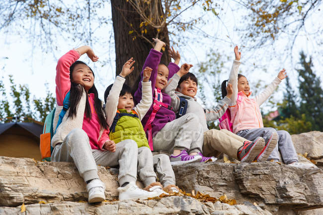 Five adorable happy asian kids sitting on stones with hands up in autumnal park — Stock Photo