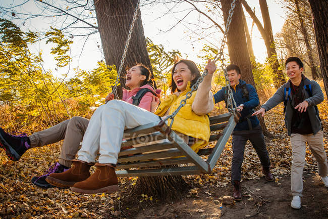 Asian men pushing happy women on swing in autumnal forest — Stock Photo