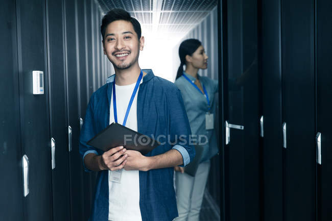 Trabajador masculino sonriendo a la cámara mientras una ingeniera trabaja en la inspección de la sala de mantenimiento - foto de stock
