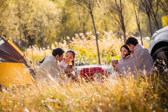 Quatro asiáticos amigos beber chá e falar no acampamento lugar no outonal floresta — Fotografia de Stock