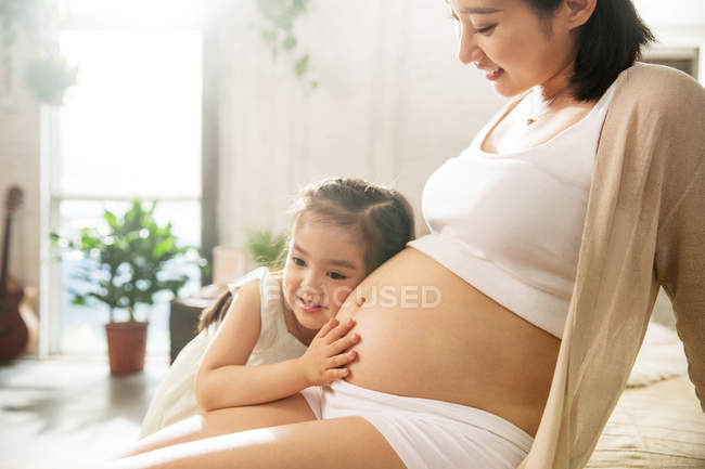 Entzückendes glückliches kleines Mädchen, das sich umarmt und dem Bauch der schwangeren Mutter zuhört — Stockfoto