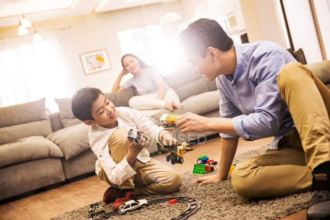 Счастливый азиатский отец и сын играют с игрушками на ковре, мать сидит на диване позади — стоковое фото