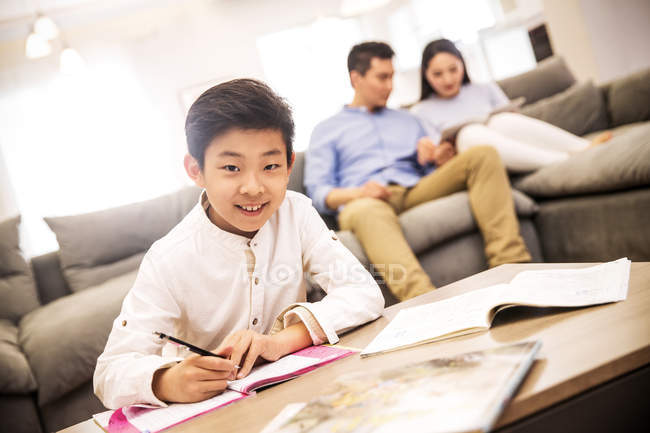 Chinese boy doing homework and smiling at camera while parents sitting on sofa behind — Stock Photo