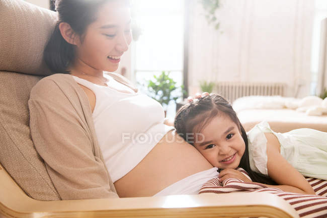 Мила усміхнена маленька дівчинка слухає живіт щасливої вагітної матері вдома — стокове фото