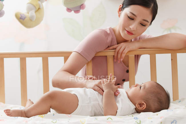 Schöne junge asiatische Frau schaut auf entzückendes Baby, das in der Krippe liegt — Stockfoto