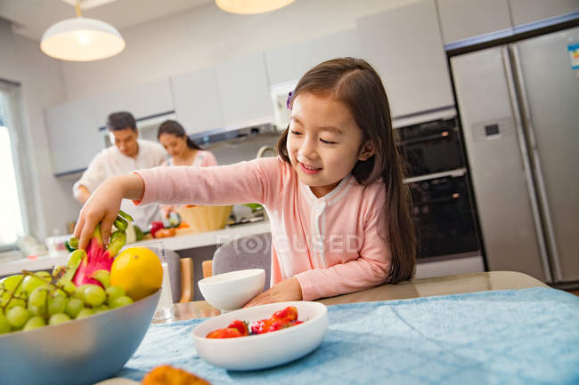 Очаровательный ребенок ест фрукты, в то время как родители приготовления пищи позади на кухне — стоковое фото