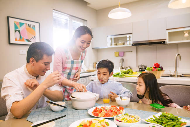 Счастливая мама приносит блюдо для улыбающейся семьи, сидящей за столом дома — стоковое фото