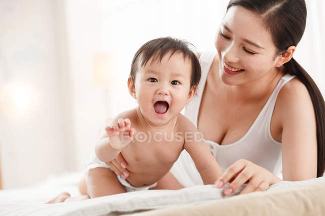 Happy young mother looking at adorable laughing baby in diaper on bed — Stock Photo