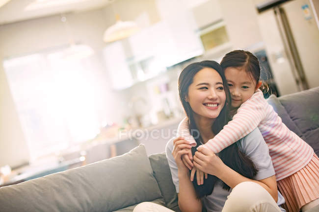 Счастливая молодая азиатская мать с очаровательной маленькой дочерью, обнимающей друг друга на диване — стоковое фото