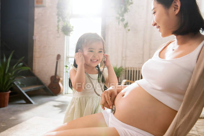 Милая улыбающаяся дочь держит стетоскоп и слушает живот беременной матери дома — стоковое фото