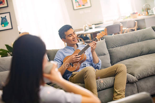 Молода жінка дивиться на чоловіка, який грає укулеле вдома — стокове фото