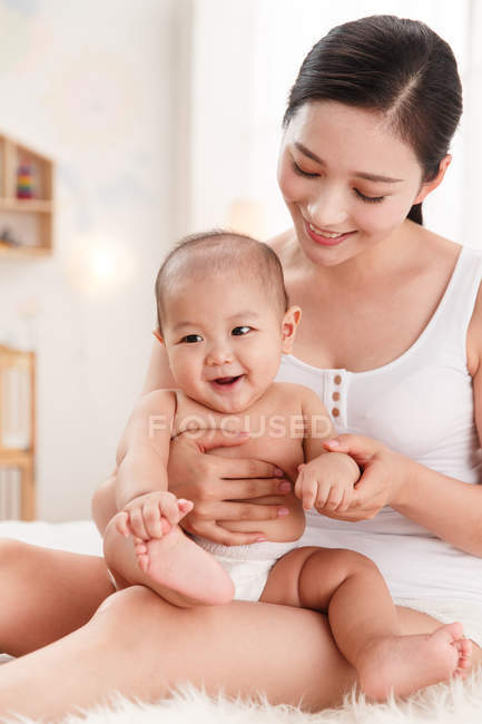 Feliz joven asiático mujer sentado con adorable bebé en la cama - foto de stock