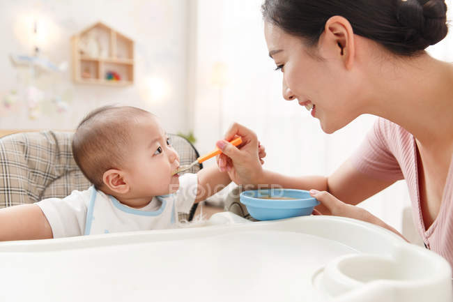 Feliz jovem mãe segurando tigela e colher, alimentando bebê adorável em casa — Fotografia de Stock