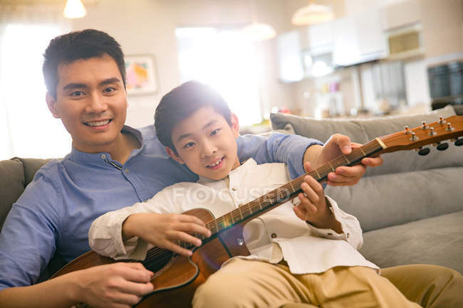 Feliz asiático pai e filho tocando violão e sorrindo para a câmera em casa — Fotografia de Stock