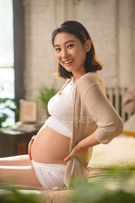 Вид сбоку счастливой молодой беременной женщины, сидящей на кровати и улыбающейся перед камерой — стоковое фото