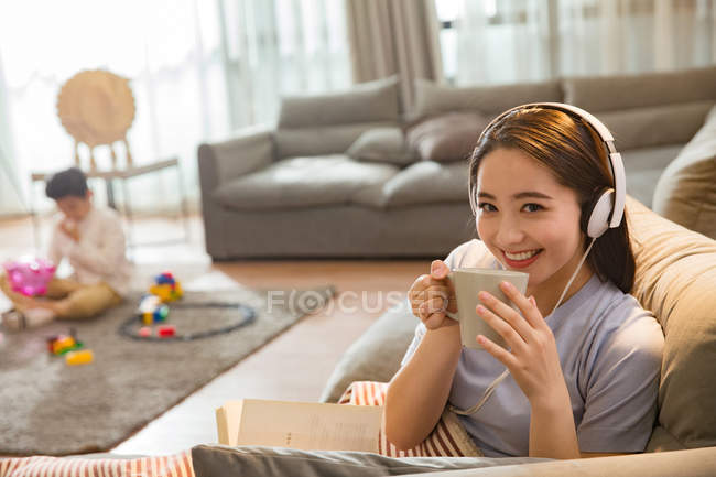 Mulher chinesa em fones de ouvido segurando copo e sorrindo para a câmera enquanto filho brincando com brinquedos atrás em casa — Fotografia de Stock