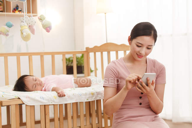 Sorrindo jovem asiático mulher usando smartphone enquanto bebê dormindo no berço — Fotografia de Stock