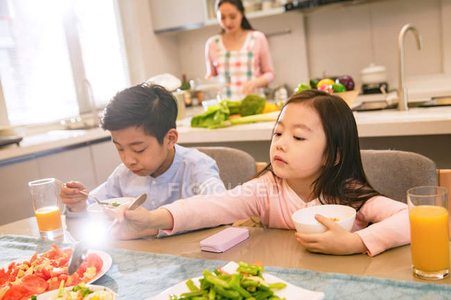 Очаровательный азиатский брат и сестра завтракают вместе, в то время как мать готовится позади на кухне — стоковое фото