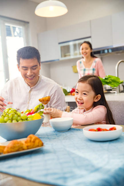 Pai com adorável sorrindo filha à mesa com café da manhã, mãe cozinhar atrás na cozinha — Fotografia de Stock