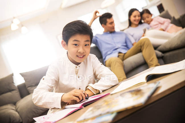 Восхитительный счастливый мальчик делает домашнюю работу и улыбается на камеру, родители с дочкой сидят сзади — стоковое фото
