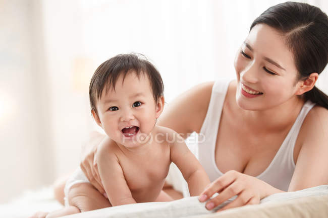 Feliz jovem mãe olhando para o bebê adorável em fralda rindo e olhando para a câmera — Fotografia de Stock