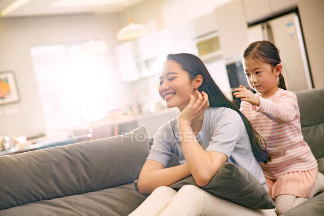 Adorable little daughter plaiting braid to smiling young mother sitting on couch — Stock Photo
