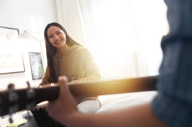 Обрезанный снимок улыбающейся молодой женщины, смотрящей на парня, играющего дома на гитаре — стоковое фото