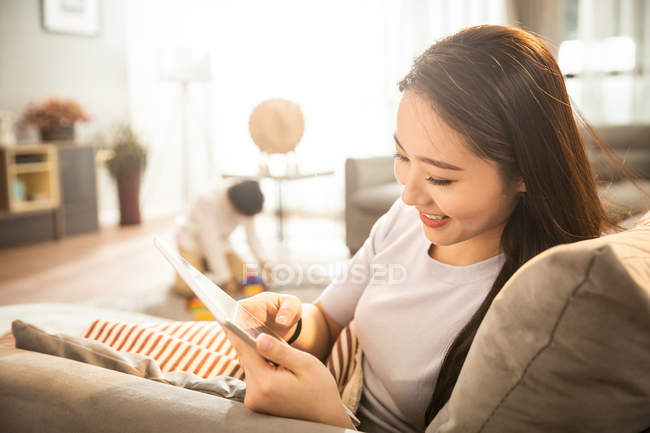 Vista lateral de mujer sonriente usando tableta digital mientras hijo jugando con juguetes detrás en casa - foto de stock