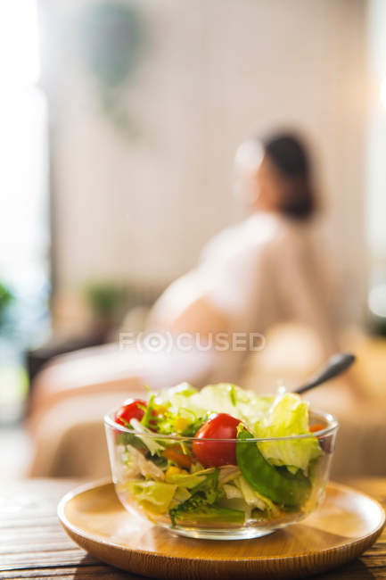 Vista de cerca del tazón con ensalada de verduras saludables y mujer embarazada sentada detrás, enfoque selectivo - foto de stock