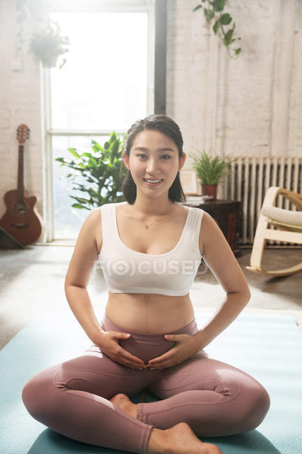 Щаслива молода азіатська вагітна жінка в спортивному одязі, сидячи на килимку йоги і посміхаючись на камеру — стокове фото