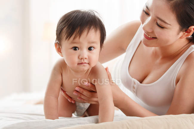 Glückliche junge Mutter schaut auf niedliches Baby, das auf dem Bett kriecht und in die Kamera schaut — Stockfoto