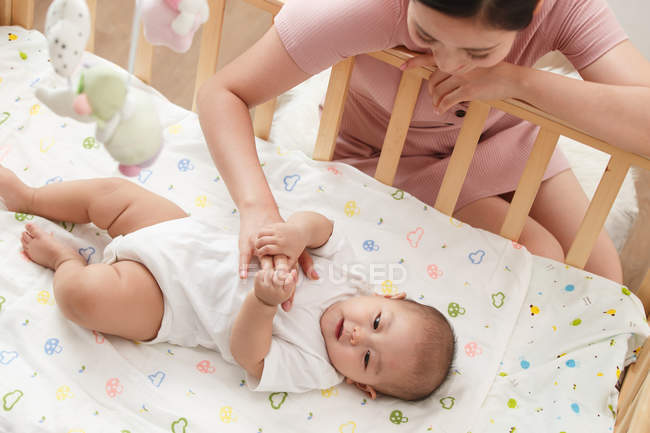 Vue grand angle de femme asiatique heureuse regardant bébé adorable couché dans la crèche, plan recadré — Photo de stock