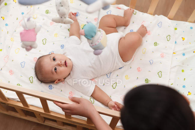 Vista aérea de la madre mirando a su adorable bebé acostado en la cama y mirando a los juguetes - foto de stock